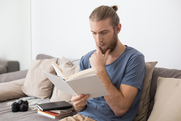 Portrait of young thoughtful man sitting on big gray sofa and reading book at home isolated