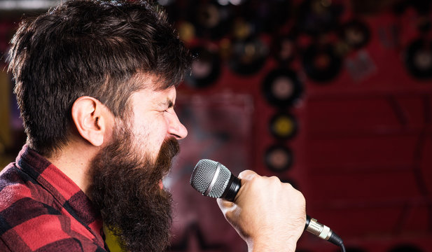 Musician with beard and mustache singing song in karaoke. Punk rock concept. Guy likes to sing in aggressive manner. Man with tense face holds microphone, singing song, club background, side view.