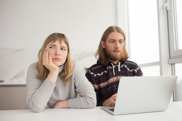 Portrait of young upset woman sadly looking aside while young man sitting near and working on laptop at home isolated