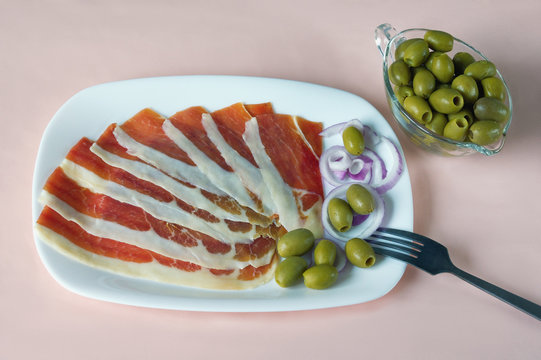 Balkan cuisine . White plate with slices of prsut (dry-cured ham,  prosciutto)  and green olives on pink pastel background