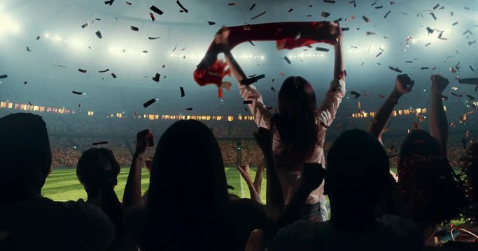 Group of cheering fans watch a sport championship on stadium. Their team wins and everybody are celebrating this event. People are dressed in casual cloth. Colourful confetti flies int the air.