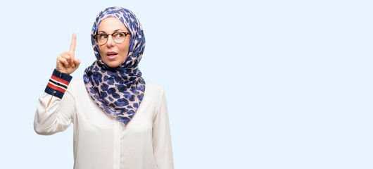 Middle age muslim arab woman wearing hijab happy and surprised cheering expressing wow gesture pointing up isolated blue background