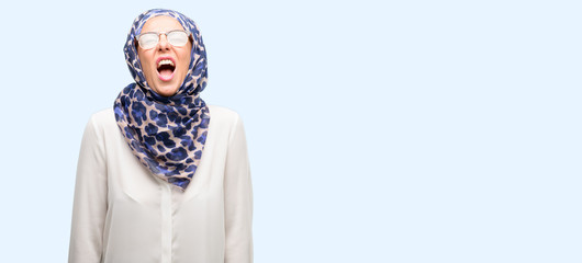 Middle age muslim arab woman wearing hijab stressful, terrified in panic, shouting exasperated and frustrated. Unpleasant gesture. Annoying work drives me crazy isolated blue background
