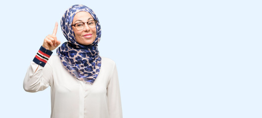 Middle age muslim arab woman wearing hijab raising finger, the number one isolated blue background