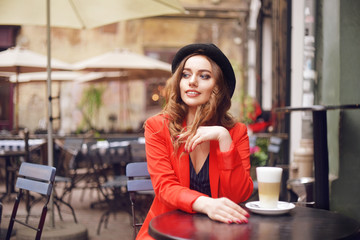 Young stylish beautiful woman sitting in city cafe in red jacket, street style, drinking aromatic coffee. Elegant girl in hat smile. Sweet breakfast, happy face, outdoor hipster portrait, fashion girl