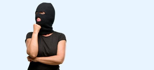 Burglar terrorist woman wearing balaclava ski mask thinking and looking up expressing doubt and wonder isolated blue background