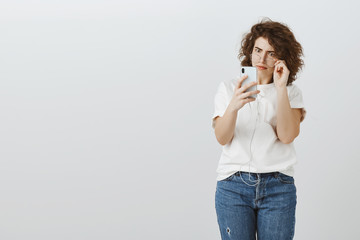 Received message shocked girlfriend. Shot of amazed curly-haired european woman, taking off glasses, holding smartphone close to face, being surprised and bothered seeing revelation over gray wall