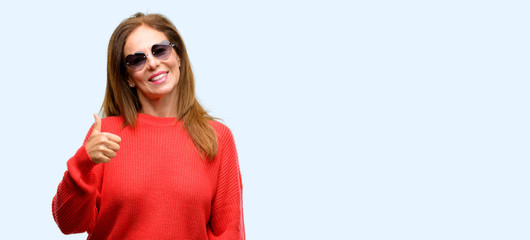 Middle age woman wearing heart sunglasses smiling broadly showing thumbs up gesture to camera, expression of like and approval isolated blue background