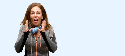 Middle age gym fit woman with workout headphones stressful keeping hands on head, terrified in panic, shouting isolated blue background