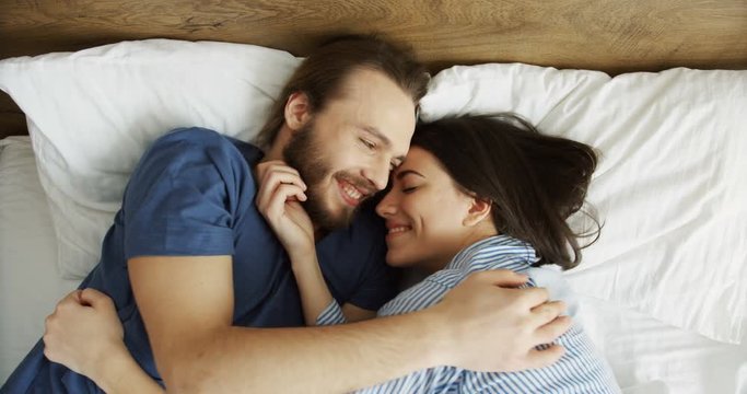 View from above on the young attractive happy man and woman lying on the bed closely, smiling and hugging. Just-married couple. Indoor