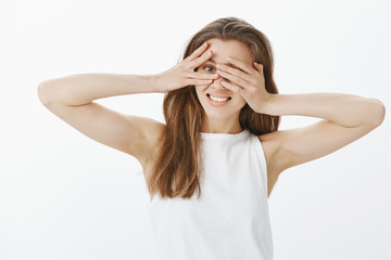 Waist-up shot of positive charming european woman with fair hair, covering eyes with palms, peeking through fingers at camera, smiling broadly, waiting for surprise or interesting gift over gray wall
