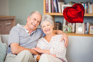 Red heart balloon against portrait of happy senior couple sitting on sofa 3d