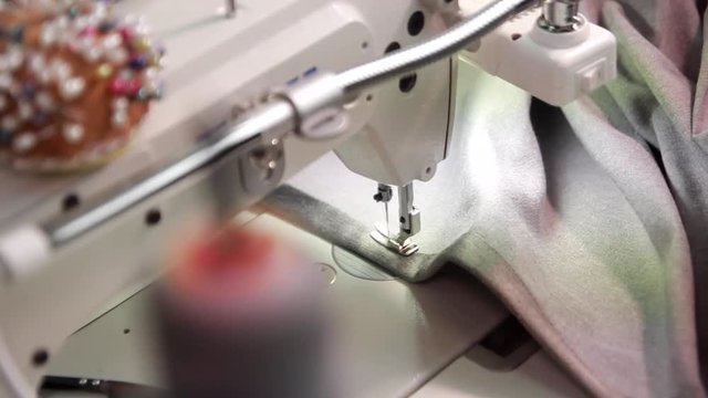Skilled seamstress work with a grey cloth on professional sewing machine under artificial lightening. A spool of high quality grey cotton thread on a sewing machine spindle on foreground, close-up. 
