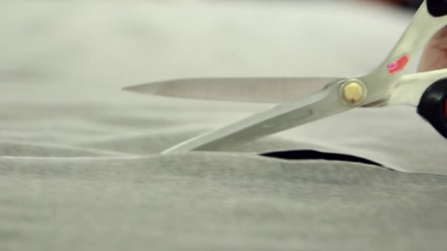 Skilled seamstress cut a grey cloth with big professional scissors. Fabric texture, close-up. Female hands in knitted sweater prepare materials with sewing tools at textile manufacture.