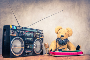 Retro Teddy Bear toy with headphones at the DJ turnable mixing console, classic mic, old cassette ghetto blaster radio recorder from 80s front concrete wall background. Vintage style filtered photo