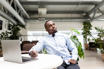 Pensive African man sitting in the office at the table making notes in a notebook