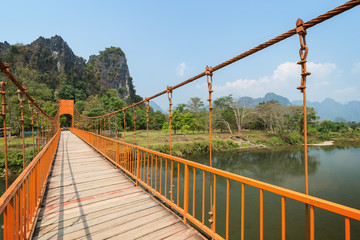 Orange suspension footbridge over the Nam Song River and scenic limestone karst mountains near the Tham Chang (or Jang or Jung) Cave in Vang Vieng, Laos, on a sunny day. - obrazy, fototapety, plakaty