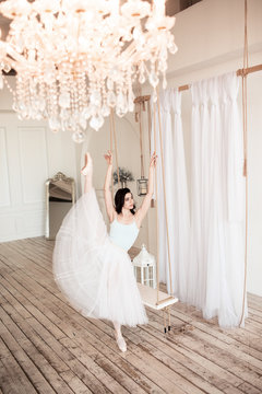 Young pretty ballerina is wearing white skirt dancing near swing in studio with light interior and wooden floor