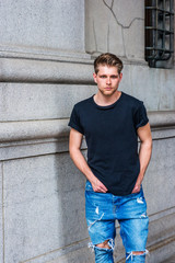 Fototapeta premium Young Modern American Man with little beard thinking outside in New York, wearing black T shirt, destroyed jeans, fashionable shoes, sitting on metal pillar on vintage street, relaxing..