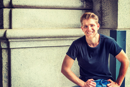 Young Handsome American College Student With Little Beard, Wearing Black T Shirt, Sitting On Street In New York, Looking At You, Smiling..