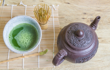 Japanese matcha green tea in stone bowl, brown clay teapot, and bamboo whisk on a bamboo mat as seen from above. Together they are the pieces of a Japanese tea ceremony