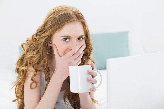 Shocked blond drinking coffee while using laptop in bed
