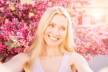 Young beautiful woman traveler making selfie photo with smart phone while walking in the street in a sunny day over pink blossom trees in city center. Close up portrait.