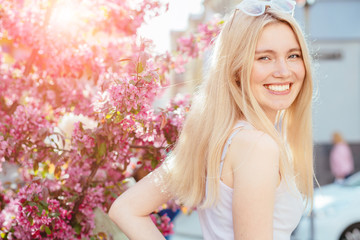 Obraz na płótnie Canvas Close up portrait of sensitive blond woman is turn around at camera and smiles. She has great smile and wonderful big blue eyes. She is playfully looks at the camera. Background pink blooming park.