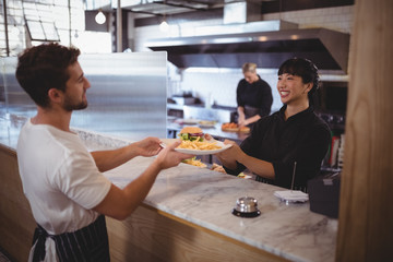 Smiling young female chef giving burger and fries to waiter