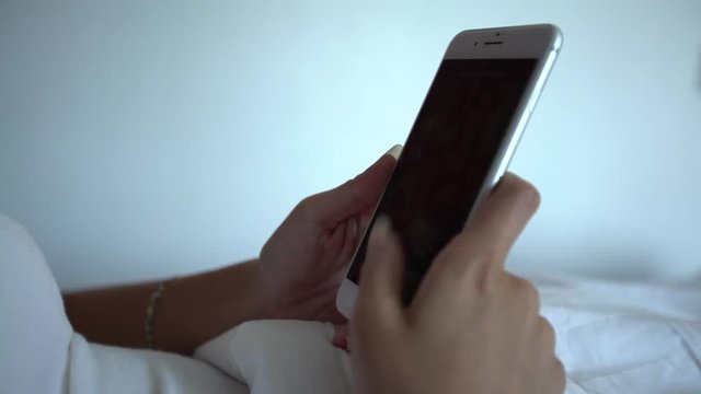 4K Asian woman lying on the bed with white sheets and using smartphone in a beautiful hotel room. Surfing the internet, checks social network and see pictures on her phone device. -Dan