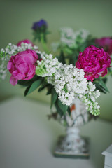 A charming bouquet of purple peonies and white lilacs in a beautiful vase on a green wall background in the living room. Selective focus.