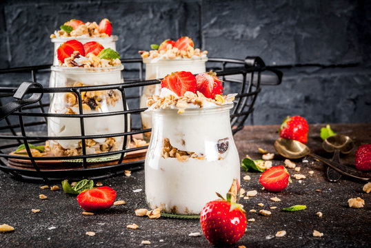 Healthy summer breakfast idea, homemade layered parfe dessert in small jar with granola, yoghurt and strawberry, dark background copy space
