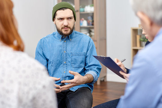 Depressed bearded man wearing denim shirt discussing his phobia with highly professional psychologist and other patients while participating in group therapy session