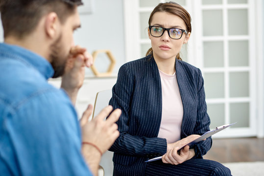 Depressed bearded man having face to face problem discussion with talented psychologist while sitting at cozy office, she holding clipboard in hands and listening to him with concentration