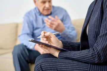 Close-up shot of female psychologist wearing elegant suit taking necessary notes on clipboard while consulting senior patient at modern office