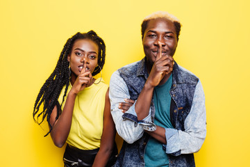 Portrait of a smiling young afro american couple showing silence gesture isolated over yellow background