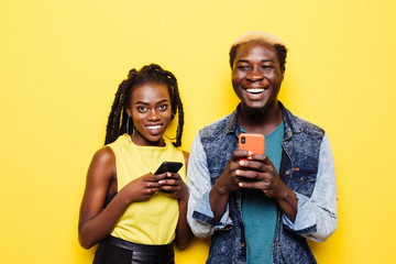Portrait of an excited young afro american couple holding mobile phones isolated on yellow...