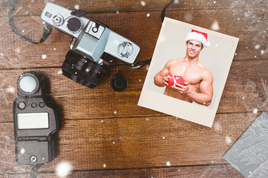 Shirtless macho man in santa hat holding gift against view of an old camera with photo flash