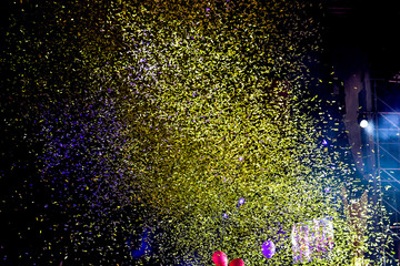 Hundreds of confetti fired on air during a festival at night. Image ideal for backgrounds. Green/yellow tonality. People with the hands to the sky. Balloons and stage on the right
