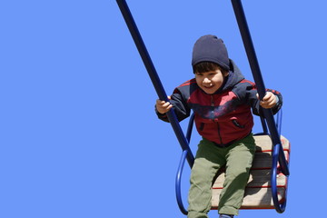 Little cute brown-eyed brunette toddler boy with pleasure swinging on a swing, smiling, laughing, blue sky background