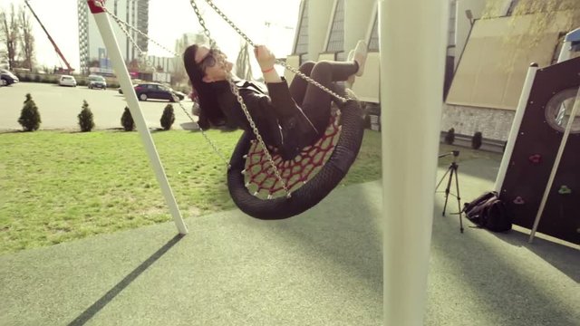 Joyful pretty girl riding on a swing in a black leather jacket and white sneakers. Swing on a Sunny day.