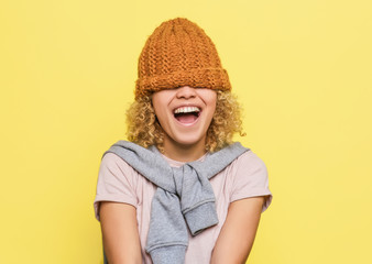 Happy and positive girl has put the brown hat onto her eyes. She can't see anything. But she is satisfied. Isolated on yellow background.