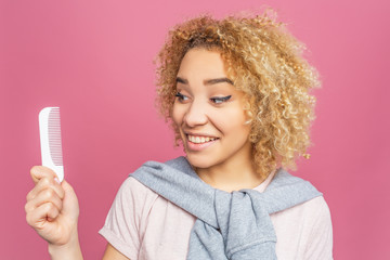 Pretty girl is looking to the brush that she holds in her hands and smiling. She cares about her blonde and curly hair. Isolated on pink background.