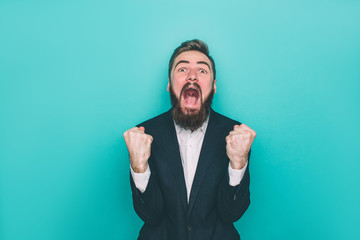 Man is screaming and yelling. He is absolutly happy. Guy is looking to camera and holding his fingers in fist. Isolated on blue background.