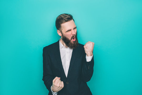 Handsome man is sreaming because of happiness. He is holding his fingers in a fist. Isolated on blue background.