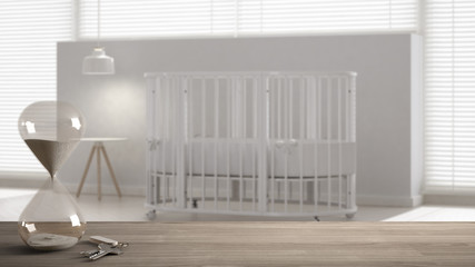 Wooden table, desk or shelf with crystal modern hourglass measuring the passing time in a countdown to a deadline and house keys over blurred nursery, architecture interior design, copy space 