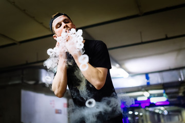 Man in cap smoke an electronic cigarette and releases clouds of vapor performing various kind of vaping tricks