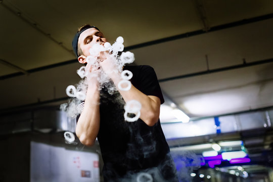 Man in cap smoke an electronic cigarette and releases clouds of vapor performing various kind of vaping tricks