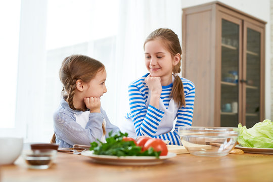 Smiling little girl wearing aprons looking at each other while discussing festive menu for Mothers Day celebration, interior of spacious kitchen on background