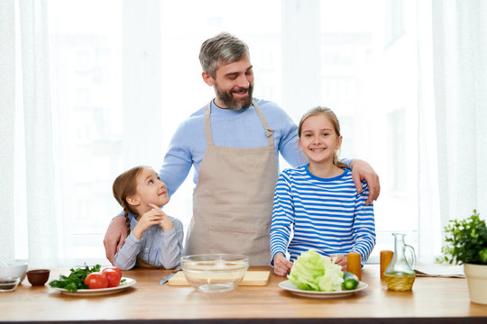 Cheerful bearded man embracing his little daughters while standing at spacious kitchen and teaching them how to cook vegetable salad, group portrait shot